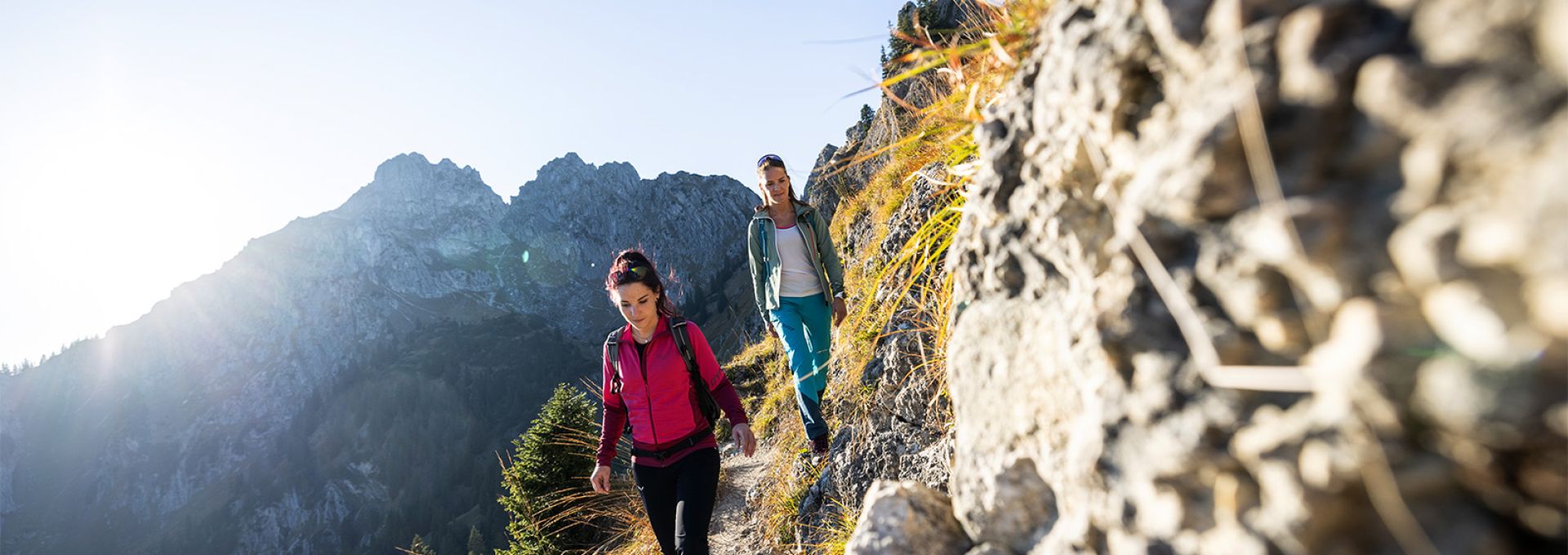 Wandern in den Ammergauer Alpen Das Bild zeigt zwei Frauen, die auf einem schmalen Bergpfad wandern. Der Pfad führt entlang eines felsigen Abhangs mit spärlicher Vegetation. Im Hintergrund erheben sich majestätische Berge, die von der Sonne angestrahlt werden. Die Szenerie ist von einer klaren, sonnigen Atmosphäre geprägt. Die Frauen sind sportlich gekleidet und wirken konzentriert auf ihren Weg.