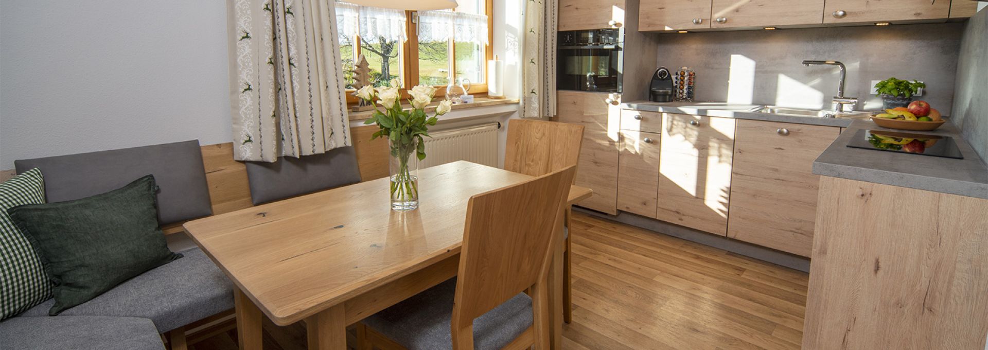 Wohnküche der Ferienwohnung Hörnle im Gästehaus Alpenglühn Das Bild zeigt eine helle, moderne Küche mit integrierter Essecke. Die Küche ist in hellen Holztönen gehalten und verfügt über moderne Geräte. Ein Esstisch aus hellem Holz steht im Vordergrund, umgeben von Stühlen mit grauen Stoffbezügen. Eine Eckbank mit grauen Kissen ist ebenfalls vorhanden. Ein Fenster mit hellen Vorhängen lässt viel Tageslicht in den Raum. Auf dem Tisch steht eine Vase mit weißen Rosen. Die Atmosphäre ist freundlich und einladend.