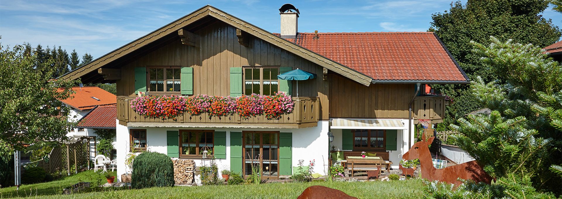 Gästehaus Alpenglühn, Bad Kohlgrub Das Bild zeigt ein zweistöckiges Haus im alpenländischen Stil mit einem roten Ziegeldach, braunen Holzwänden und grünen Fensterläden. Vor dem Haus befindet sich ein Balkon, der mit bunten Blumen geschmückt ist. Der Garten ist grün und gepflegt, mit zwei rostigen Metallfiguren von Rehen. Der Himmel ist sonnig und blau. Das Gesamtbild wirkt idyllisch und ruhig.