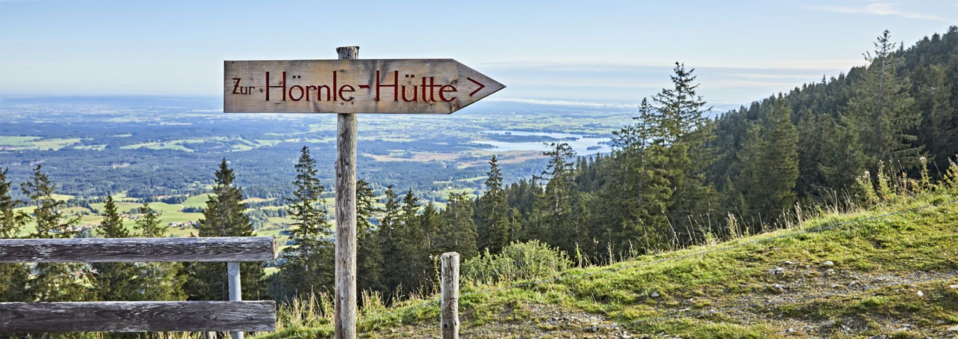 Anreise Das Bild zeigt einen hölzernen Wegweiser mit der Aufschrift „Zur Hörnle-Hütte“ an einem Berghang mit Blick auf eine weitläufige, hügelige Landschaft mit Wäldern und Seen. Im Vordergrund befindet sich ein einfacher Holzzaun. Der Himmel ist klar und blau mit ein paar wenigen Wolken. Das Bild strahlt Ruhe und die Schönheit der Natur aus, und lädt zum Wandern ein.